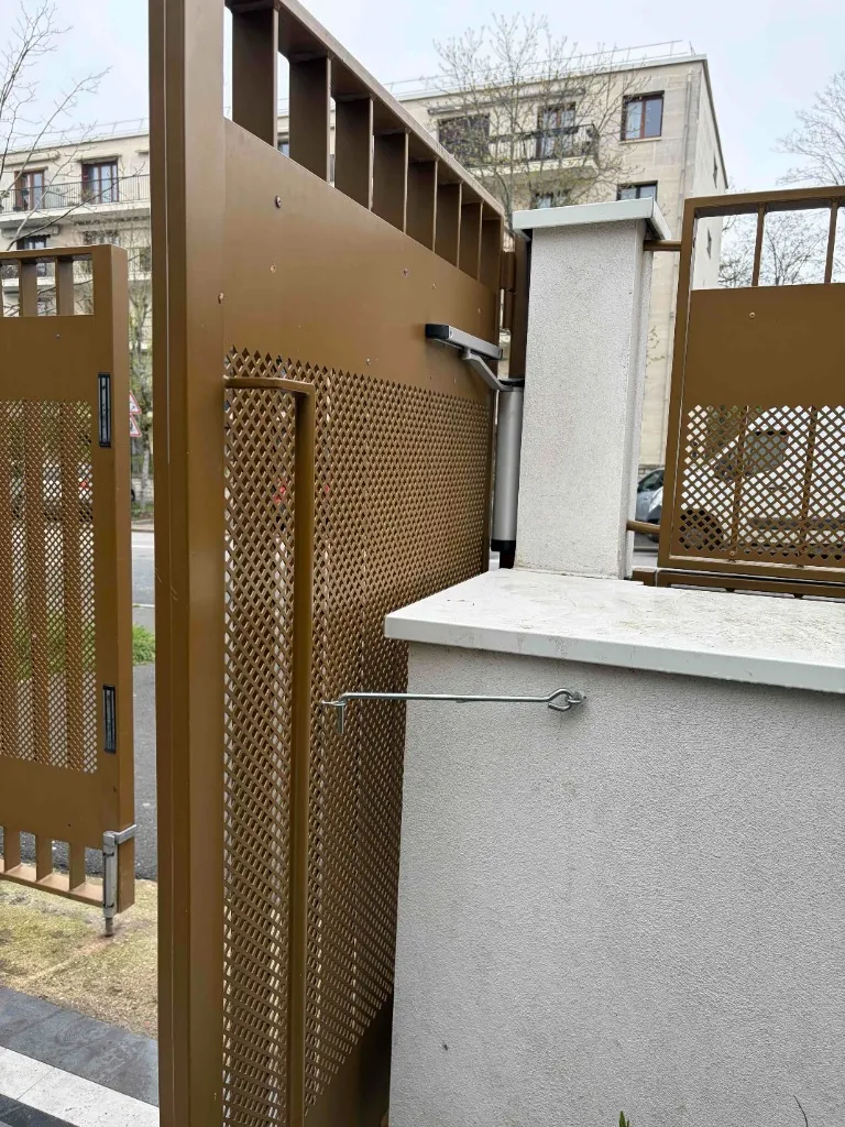 Détail d’un bras de ferme-porte mécanique fixé sur un pilier et une grille